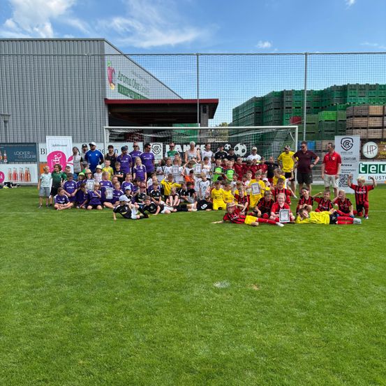 Eine große Gruppe von Kindern und Erwachsenen posiert für ein Foto auf einem Fußballfeld. Sie tragen Uniformen in verschiedenen Farben. Hinter ihnen ist ein Gebäude mit Schildern und einem Torpfosten zu sehen.
