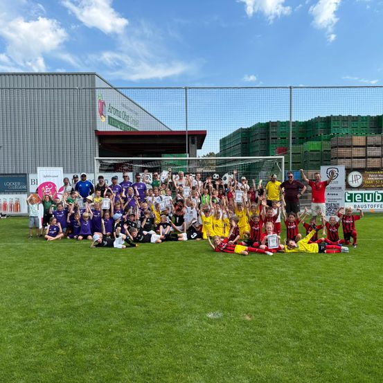 Eine Gruppe von Fußballspielern und Trainern posiert für ein Foto auf einem Feld mit einem Fußballtor im Hintergrund.
