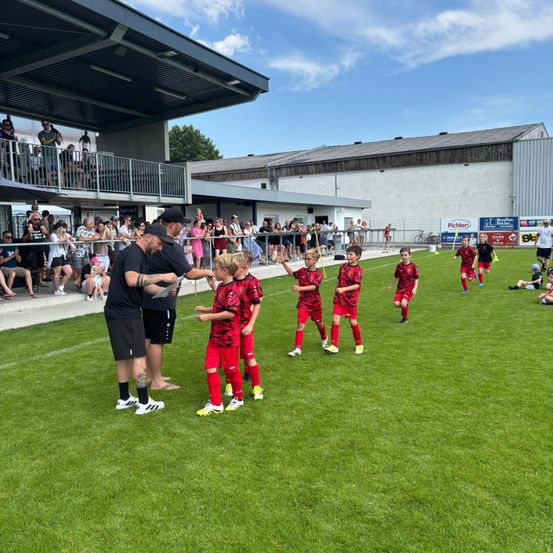 Eine Gruppe junger Fußballspieler in roten Trikots läuft auf dem Feld, begleitet von zwei Männern. Zuschauer beobachten vom Stehplatz aus.