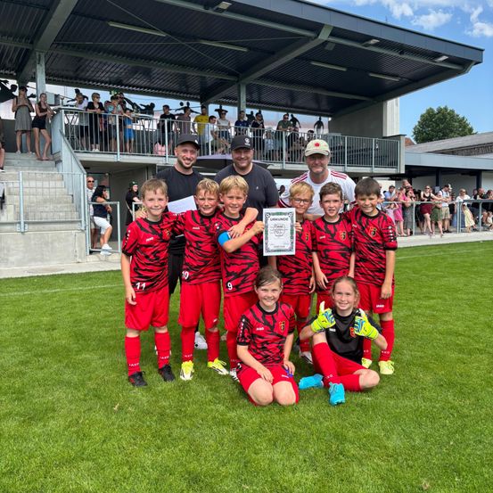 Eine Fußballmannschaft posiert mit ihrem Zertifikat auf dem Spielfeld, während einige Personen hinter ihnen auf der Tribüne stehen.