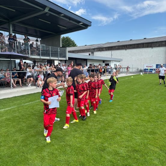 Eine junge Fußballmannschaft in roten und schwarzen Uniformen läuft über ein Feld, während Zuschauer hinter einem Zaun zusehen.