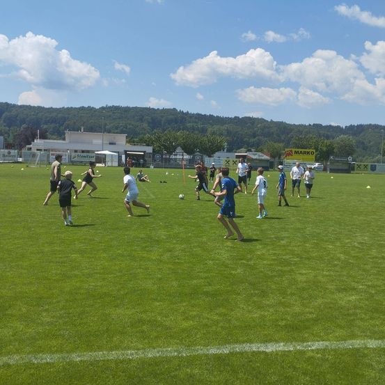 Eine Gruppe von Kindern spielt Fußball auf einem Feld unter blauem Himmel. Sie rennen mit gelben Markierungen um das Feld herum, tragen Shorts und Sneakers. Bäume und ein Gebäude sind im Hintergrund.