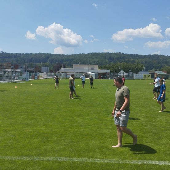 Eine Gruppe von Menschen in Shorts und T-Shirts ist auf einem grünen Feld. Ein Mann in einem grünen Shirt geht vorwärts, während andere im Hintergrund stehen. Es gibt orangefarbene Hütchen auf dem Feld und ein Gebäude in der Ferne.