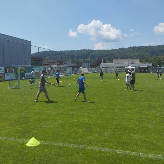 Eine Fußballtrainingseinheit mit Spielern in blauen und weißen Trikots findet auf einem grünen Rasen statt. Ein gelber Kegel ist auf der linken Seite platziert.