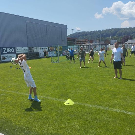 Eine Gruppe von Menschen spielt Fußball auf einem Feld mit einem jungen Jungen, der einen Fußball hält. Hinter ihnen steht ein Zaun mit Werbetafeln und einem Berg in der Ferne.