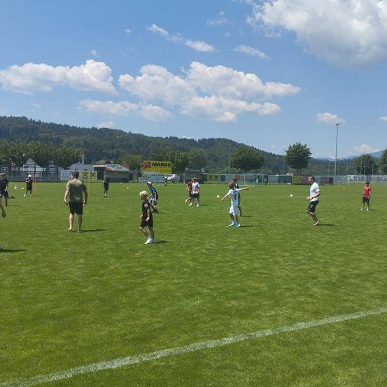 Ein Fußballtraining findet auf einem gut gepflegten Rasenplatz statt, wobei die Spieler in verschiedenen Positionen stehen und Berge im Hintergrund sind.