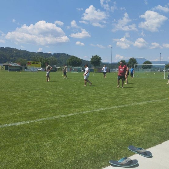 Eine Gruppe von Menschen spielt auf einem grasbewachsenen Feld mit weißen Linien, umgeben von Bergen und Bäumen. Einige sind barfuß, andere tragen Turnschuhe. Es gibt orangefarbene Hütchen und ein Fußballtor.