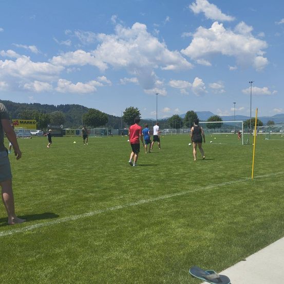 Ein Fußballfeld mit verstreuten Leuten. Einige tragen Turnschuhe, andere haben keine Schuhe. Das Feld ist breit und grün. Im Hintergrund gibt es Bäume, Berge und ein Schild.