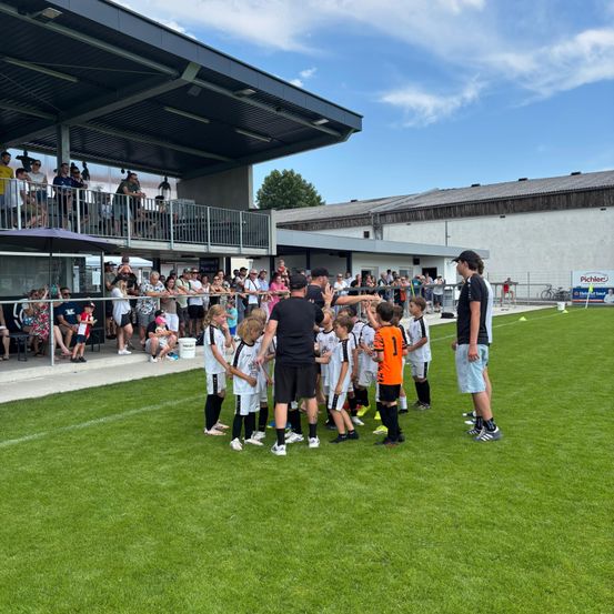 Eine Gruppe von Kindern in Fußballtrikots steht auf dem Feld, mit Trainern und Zuschauern in der Nähe. Die Umgebung ist im Freien, mit einem Gebäude und Bäumen im Hintergrund.