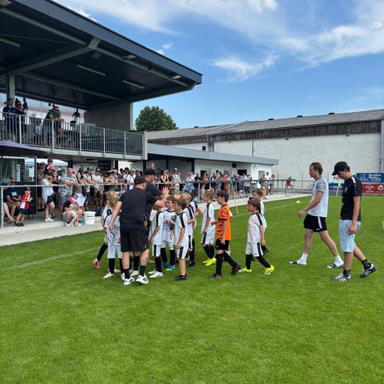 Eine Gruppe junger Fußballspieler läuft auf einem grünen Feld, begleitet von zwei Erwachsenen. Zuschauer beobachten von den Tribünen aus.