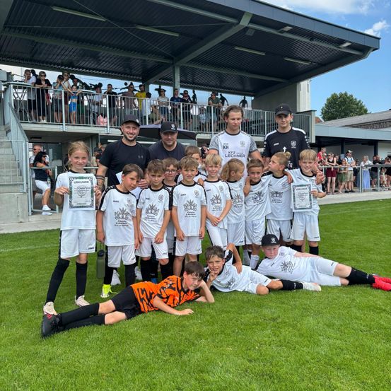 Eine Fußballmannschaft feiert mit Auszeichnungen auf dem Feld, wobei die Spieler liegen und Urkunden halten. Dahinter stehen zwei Trainer, und Zuschauer sehen von den Tribünen zu.