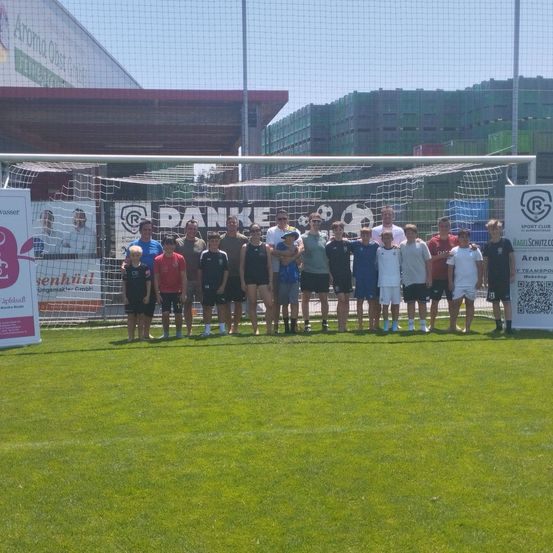 Eine Gruppe von Menschen in Sportkleidung steht vor einem Fußballtor. Sie lächeln und posieren für ein Foto. Es gibt zwei Banner an den Seiten. Der Hintergrund ist unscharf.