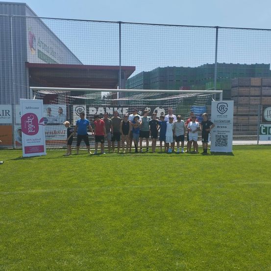 Eine Gruppe von Menschen steht auf einem Feld vor einem Tor. Dahinter befinden sich verschiedene Banner. Der Himmel ist klar, und im Hintergrund befinden sich Gebäude und ein Zaun.