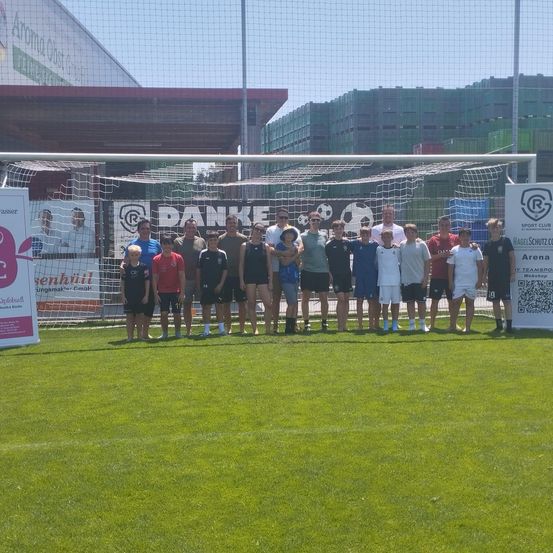 Eine Gruppe von Menschen steht vor einem Fußballtor und lächelt für ein Foto. Sie tragen verschiedenfarbige Trikots. Im Hintergrund befinden sich Gebäude, ein Netz und Werbebanner.