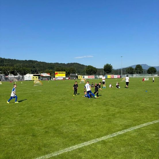 Ein Fußballfeld mit mehreren Spielern und einigen Zuschauern. Die Spieler rennen und spielen auf dem grünen Feld. Einige Spieler stehen in der Nähe der weißen Linie auf dem Feld.