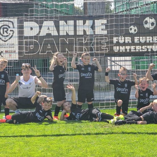 Gruppenfoto von jungen Fußballspielern in schwarzen Trikots, die hinter einem Tor posieren. Sie lächeln und zeigen ihre Fäuste in die Luft.