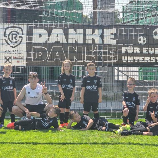 Eine Gruppe junger Fußballspieler in schwarzen Trikots mit dem Wort 'geofin' posiert für ein Foto auf einem Feld. Sie sind in der Nähe eines Netzes mit einem Banner darüber, das Danke sagt.