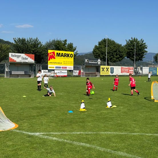 Auf einem Fußballfeld üben junge Spieler in roten Trikots mit einem Ball, begleitet von Trainingshütchen und einem Tor. Werbung ist im Hintergrund sichtbar.