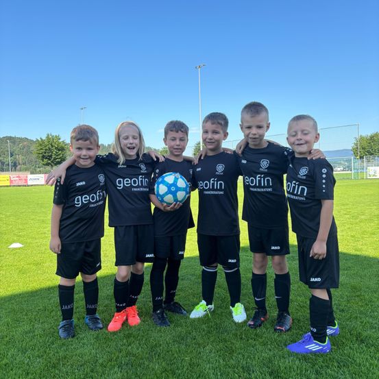 Fünf junge Fußballspieler posieren an einem sonnigen Tag für ein Foto, tragen schwarze Trikots mit dem Wort 'geofin'. Sie halten einen Fußball und lächeln für die Kamera. Dahinter ist ein Fußballfeld mit einem Tornetz.