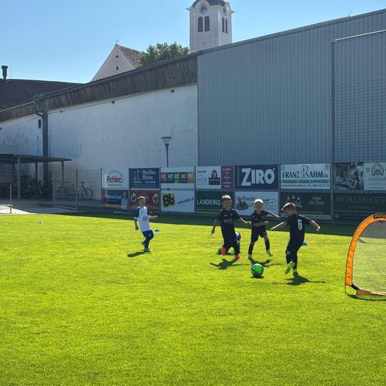 Kinder spielen Fußball auf einem grünen Feld unter einem klaren Himmel, in der Nähe eines Gebäudes mit Turm und ZIRO-Schild. In der Nähe sind Fahrräder geparkt.