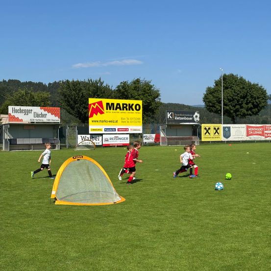 Ein Fußballtraining auf einem grünen Feld. Kinder laufen auf Fußballbälle zu. Ein gelbes Tor mit Netz steht auf dem Feld. Werbebanner und Schilder befinden sich am Zaun.