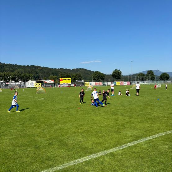 Kleine Kinder spielen Fußball auf einem Feld. Einige sind in Teams, andere stehen herum. In der Ferne sind Berge.