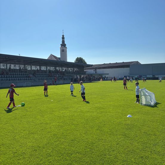 An einem sonnigen Tag spielen Kinder Fußball auf einem gut gepflegten Kunstrasenplatz mit einem Turm im Hintergrund.