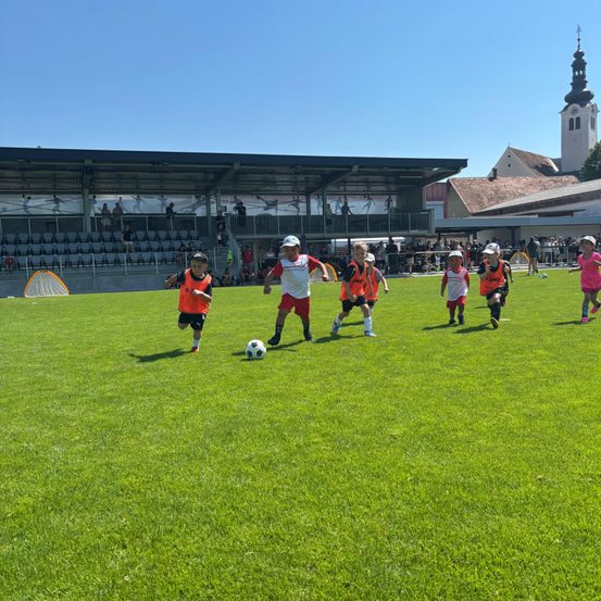 Kinder in Sportkleidung spielen Fußball auf einem Rasenfeld, mit einem Gebäude und einem Turm im Hintergrund.