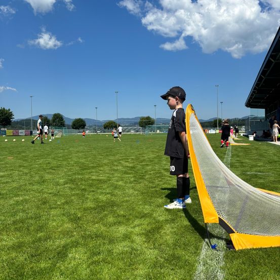 Ein Junge in einem Fußballtrikot steht neben einem Netz auf einem Feld, während andere Spieler im Hintergrund üben.