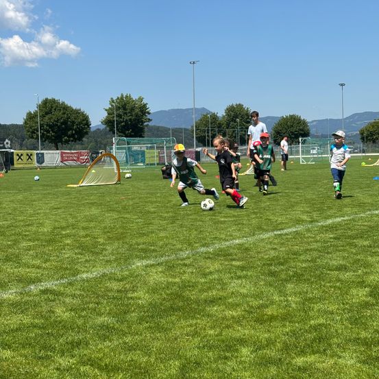 Mehrere junge Jungen spielen Fußball auf einem grünen Feld, während einige Erwachsene im Hintergrund zuschauen. Ein Junge läuft auf den Ball zu, bereit ihn zu treten.