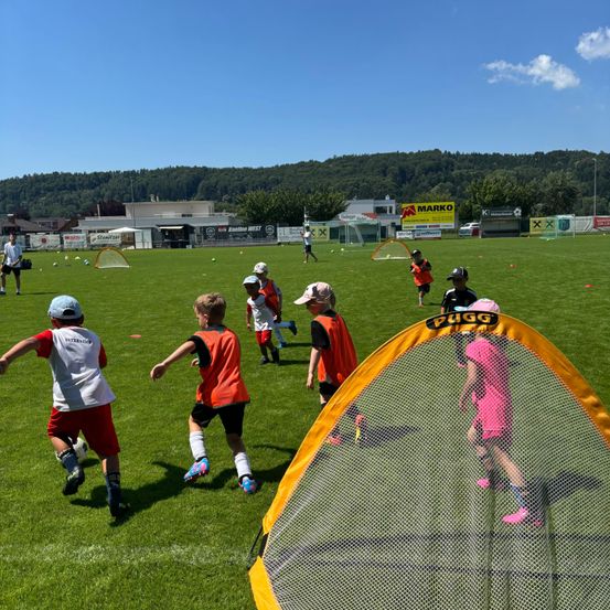 Eine Gruppe von Kindern in orangefarbenen Westen übt Fußball auf einem Feld. Vor ihnen steht ein Netz, und ein Mann steht in der Nähe. In der Ferne ist ein Fußballtor zu sehen, und mehrere Personen stehen auf dem Feld. Bäume und Gebäude sind im Hintergrund sichtbar.