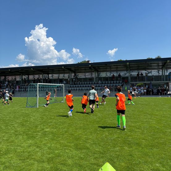 Ein Fußballspiel findet auf einem gut gepflegten Feld statt. Spieler in orangefarbenen Trikots sind über das Feld verteilt, wobei zwei Teams sichtbar sind. Zuschauer sind anwesend, einige stehen an den Seitenlinien und andere sitzen in den Rängen.