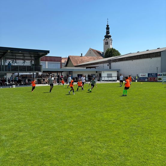 Eine Gruppe von Kindern in orangefarbenen Uniformen spielt Fußball auf einem grünen Feld unter klarem Himmel, mit einem Gebäude im Hintergrund.