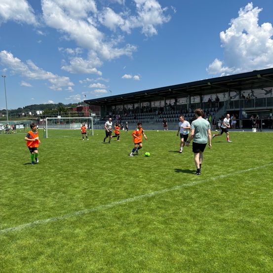 Mehrere Menschen spielen Fußball auf einem grünen Feld unter einem blauen Himmel. Erwachsene und Kinder in orangefarbenen Trikots rennen. Ein Mann in einem grünen Shirt geht.
