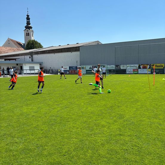 Junge Fußballspieler in orangefarbenen Trikots üben auf einem grünen Feld. Im Hintergrund steht ein Gebäude mit Turm.