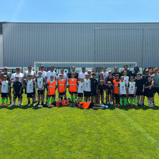 Eine Fußballmannschaft posiert für ein Gruppenfoto auf einem Rasenfeld, mit Spielern in weißen, schwarzen und orangefarbenen Trikots. Hinter ihnen steht ein Gebäude mit einer Metallwand und einem Tor.