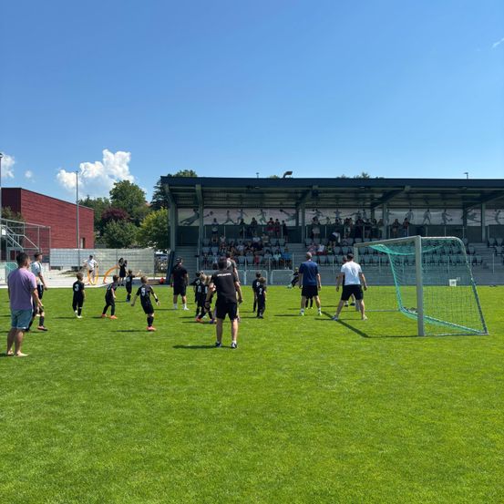 Ein Fußballtraining auf einem Feld mit Kindern in nummerierten Trikots, zwei Erwachsenen vorne, einem Tor und Zuschauern auf den Tribünen.