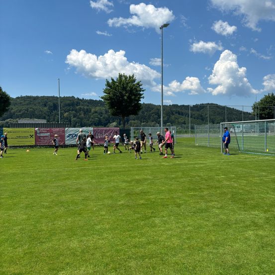Eine Gruppe von Menschen spielt Fußball auf einem gut gepflegten grünen Feld mit Bäumen und Bergen im Hintergrund.