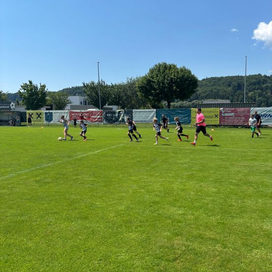 Eine Gruppe junger Spieler trainiert auf einem Feld unter klarem Himmel mit Bäumen und Bergen im Hintergrund Fußball.