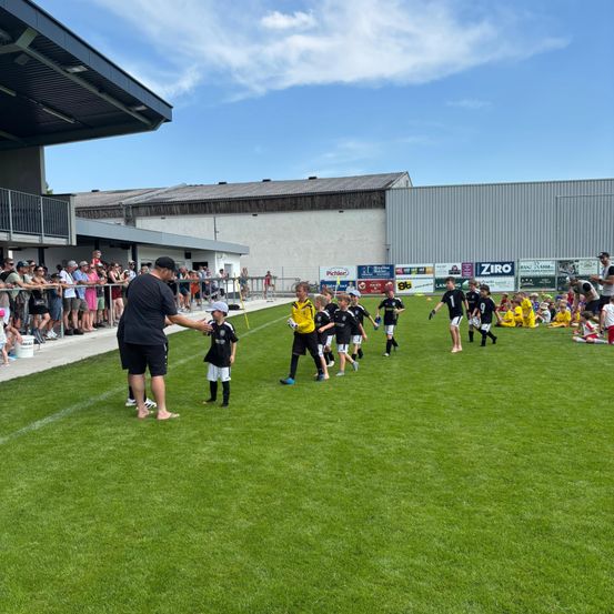 Ein Fußballtrainer führt eine Mannschaft junger Spieler auf einem Feld. Zuschauer beobachten von den Rändern. Der Himmel ist teilweise bewölkt.