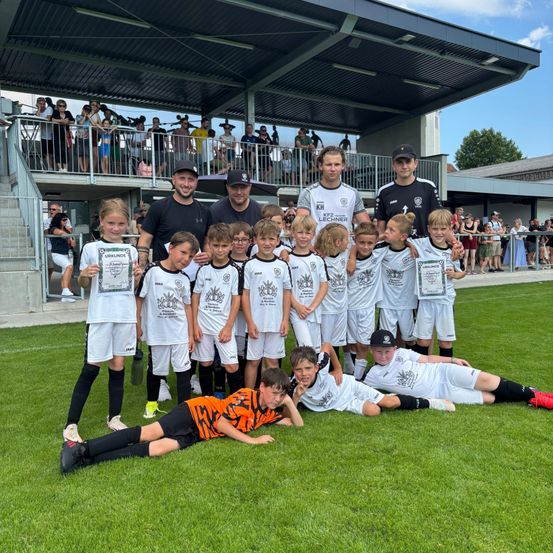 Eine Fußballmannschaft mit jungen Spielern in weißen Trikots posiert für ein Foto auf dem Feld, mit einigen Erwachsenen und Zuschauern im Hintergrund.