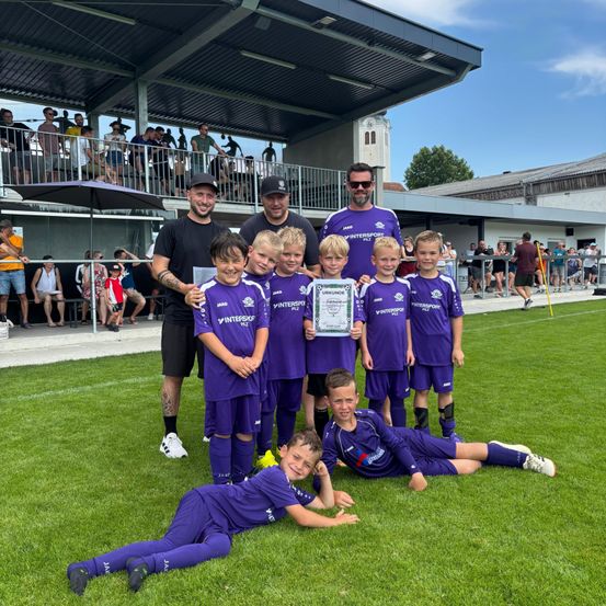 Eine Fußballmannschaft in lila Trikots posiert für ein Foto auf dem Feld. Sie halten einen Zettel mit 'Urkunde' darauf. Hinter ihnen stehen mehrere Leute.