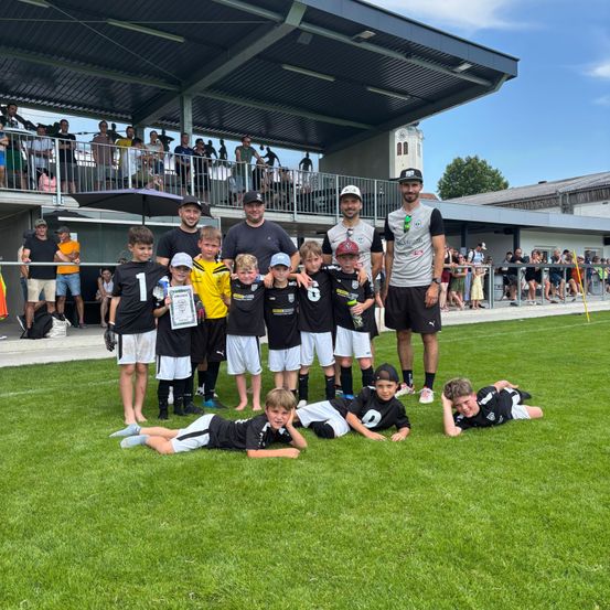 Eine Jugendfußballmannschaft posiert für ein Foto auf dem Feld, einige Spieler liegen auf dem Rasen. Erwachsene stehen hinter ihnen, und Zuschauer sind im Hintergrund.