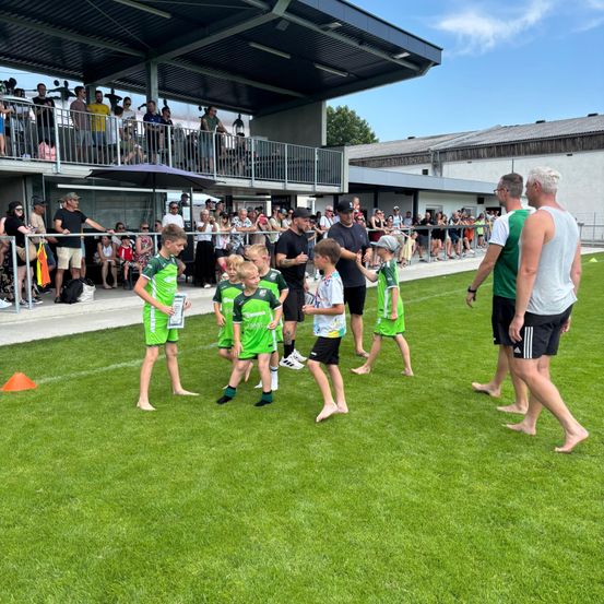 Mehrere junge Jungen in grünen Trikots stehen auf einem grünen Feld, wahrscheinlich bei einer Sportveranstaltung. Zuschauer sehen von den Tribünen zu. Erwachsene stehen in der Nähe.