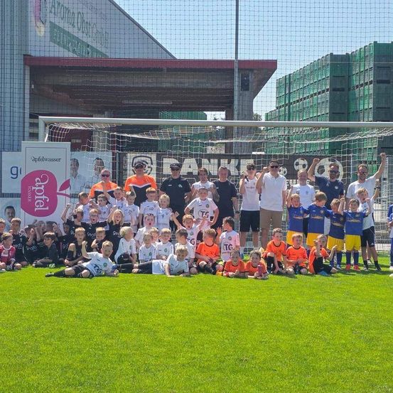 Eine Gruppe von Kindern und Erwachsenen posiert für ein Foto auf einem Fußballfeld mit einem Tor. Viele tragen orangefarbene und weiße Trikots.