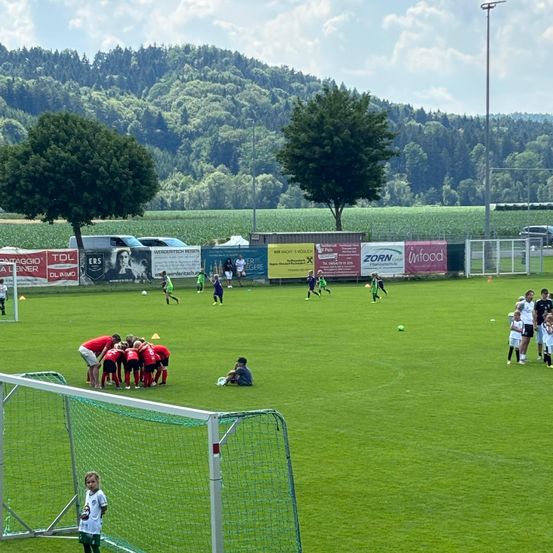 Fußballspieler versammeln sich auf einem Feld vor dem Hintergrund von Bergen. Einige üben mit Bällen, während andere in der Nähe eines Tores stehen. Bäume und Banner säumen die Seiten des Feldes.