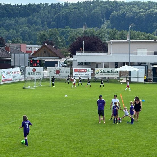 Ein Fußballplatz mit spielenden Kindern. Mehrere Spieler sind auf dem Feld, und einige stehen in der Nähe des Tores. Das Feld ist von Gebäuden umgeben, und eine Anzeigetafel befindet sich links.