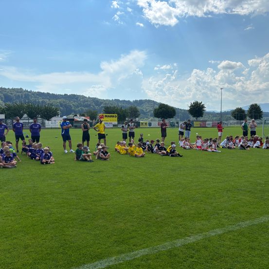 Eine Fußballmannschaft versammelt sich auf einem Feld mit Spielern in blauen und gelben Trikots, Trainern und Zuschauern. Der Himmel ist teilweise bewölkt, und Berge bilden den Hintergrund.