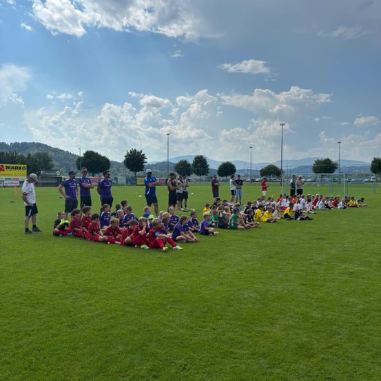Eine Gruppe junger Fußballspieler sitzt auf dem Feld, umgeben von Trainern, während andere stehen und zusehen unter einem bewölkten Himmel.