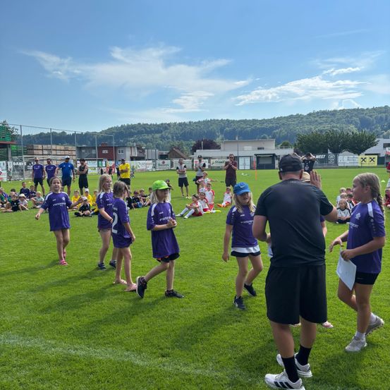 Kinder in lila Uniformen laufen auf einem grünen Feld. Ein Mann in Schwarz gibt ihnen einen High Five. Dahinter sitzen Leute auf dem Gras und beobachten sie. In der Ferne gibt es Gebäude und Bäume.